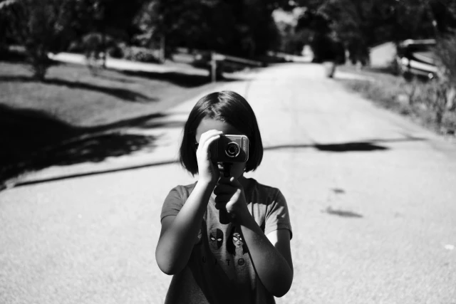 A blond girl holds a camera to her face, obscuring it completely, facing the photographer. She stands in front of an empty road with a long tall shadow behind her.