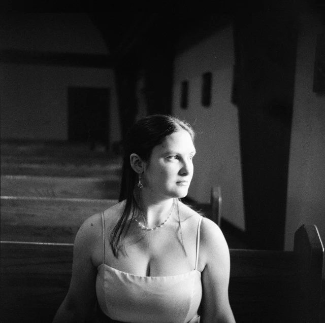 A brown-haired woman in a spaghetti strap dress looks into thin light inside a dim church.